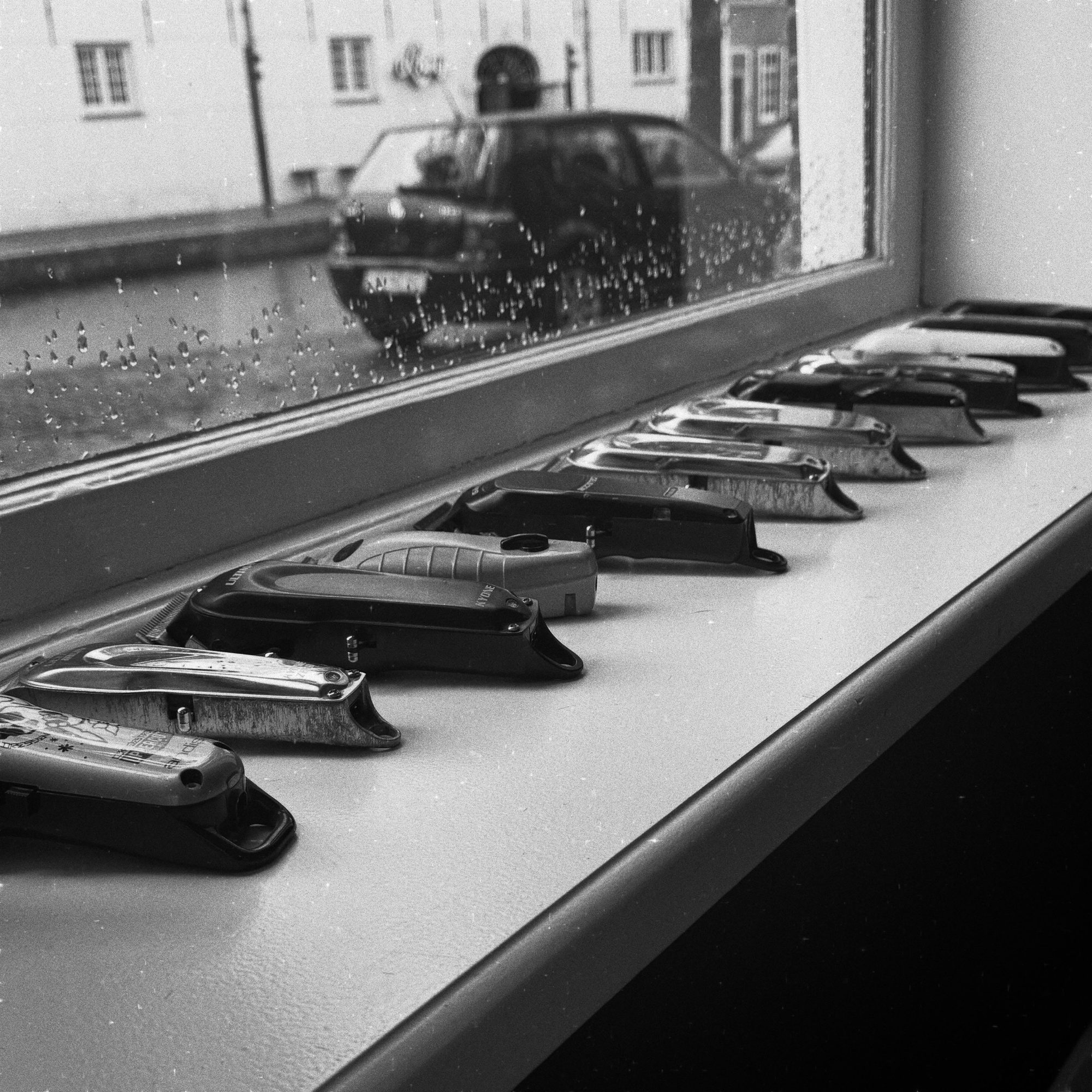 Een rij tondeuses op de vensterbank van de shop, met regen op het raam en de Lange Geer erachter