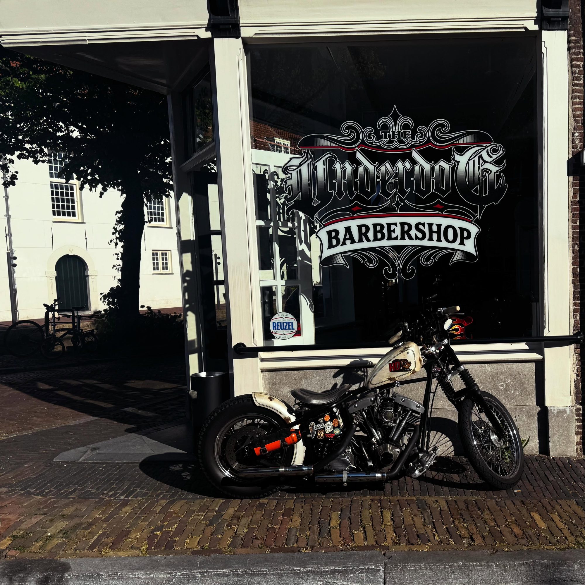 De gevel van The Underdog Barbershop aan de Lange Geer, met het logo op het zwarte raam en een motor ervoor
